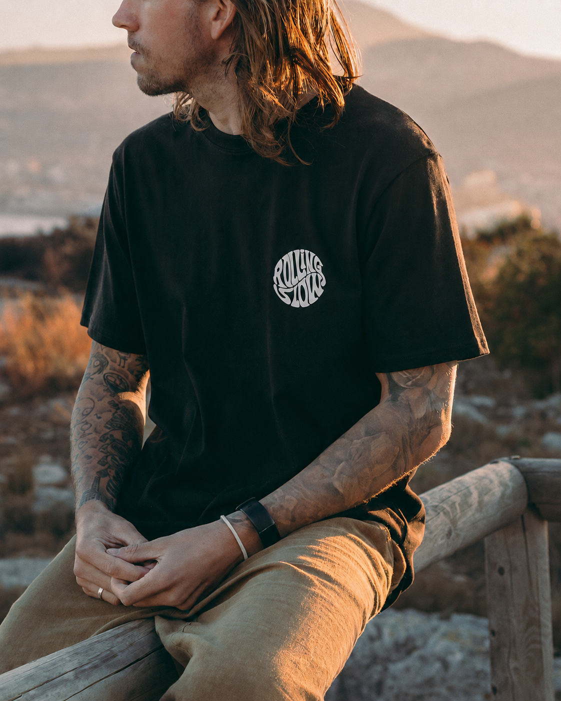 Person wearing a black t-shirt with a logo, sitting outdoors with mountains in the background